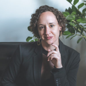 Bethany with short curly hair and her hand resting on her chin wearing a black suit jacket sitting on a couch in front of a light colored wall and a green plant behind her.