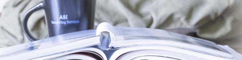 Image of a binder and books sitting on a table with an ASL Interpreting mug sitting behind them.