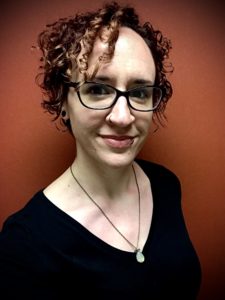 Image of Bethany wearing glasses and a long necklace with a slight smirk standing in front of a coral colored wall.