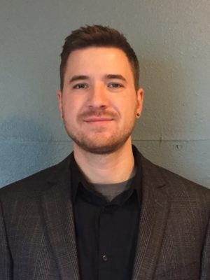 Image of Henry Yandrasits standing in front of a gray wall wearing a gray suit jacket and black button up smiling at the camera.