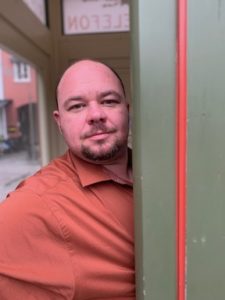 Image of a white male leaning against a wall with a slight smirk. 