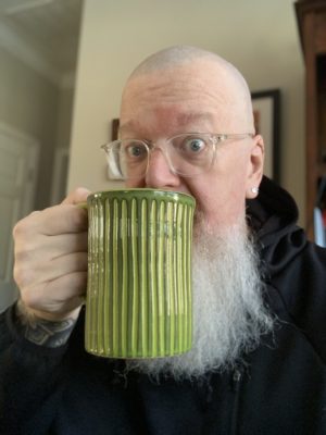 White bald male with a white beard wearing light colored glasses and a black hoodie with a green coffee mug to his lips.