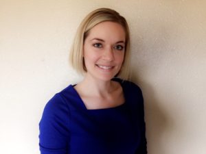 image of Jana standing in front of a cream colored wall wearing a blue shirt and smiling at the camera