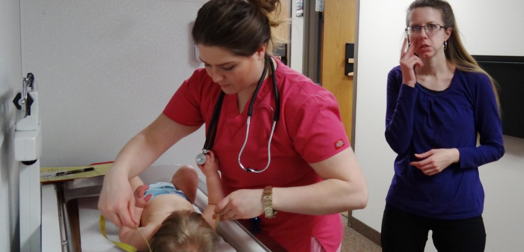 Image of a nurse performing a regular check-up on an infant. Standing behind the nurse is an interpreter signing "SEE".