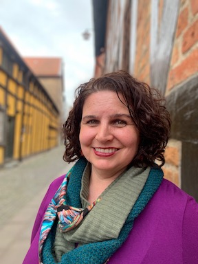 Image of Naomi Sheneman standing against a brick wall smiling at the camera.