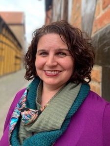 Image of woman smiling at camera with curly short hair leaning against a brick wall. 