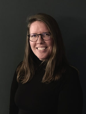 Paula Clark wearing black rimmed glasses and a black turtle neck sitting in front of a black background.