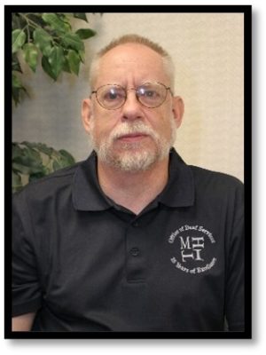Image of a white male wearing glasses in front of a tan wall. He is wearing a black collared shirt with the MHIT logo on the right side that reads: Office of Deaf Services 25 Years of Excellence.