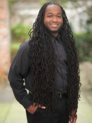 Image of Vyron Kinson standing outside wearing a grey button up and black dress pants with his hand in his front pocket, smiling at the camera.