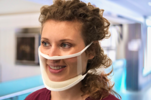 Woman with curly shoulder length hair wearing the "clear mask". This mask is transparent and shows her entire face. There is a chin padding and piece that sets just slightly above the peak of her nose.