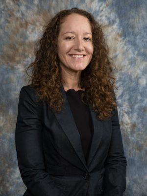 Jasmine, a white female with long curly dark hair, wearing a black shirt and dark grey suit jacket standing smiling.