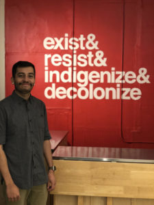 Image of a latino man smiling. The background behind him is a read wall with white letters that reads, "exist&resist&indigenize&decolonize"